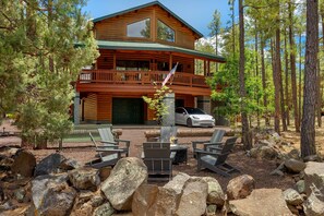 A two-story log cabin with a balcony, a stone fireplace, and a white car parked in front. - RainbowLake-HotTub-GameRoom-Kayaks-Horseshoes-EVCharge-Firepit-Pickleball-7Beds (Pinetop-Lakeside)