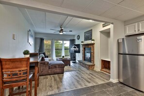 A modern kitchen with a built-in refrigerator, a dining area with a wooden table and chairs, a fireplace, and a large window with a view of greenery. - Treetop Hearth at Mountain Green | 1BR Balcony Condo | Sleeps 4 (Killington)