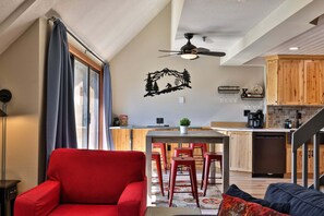 En moderne stue med en rød sofa, et spiseområde med et træbord og røde barstole, samt et køkken med træskabe og et sort køleskab.
