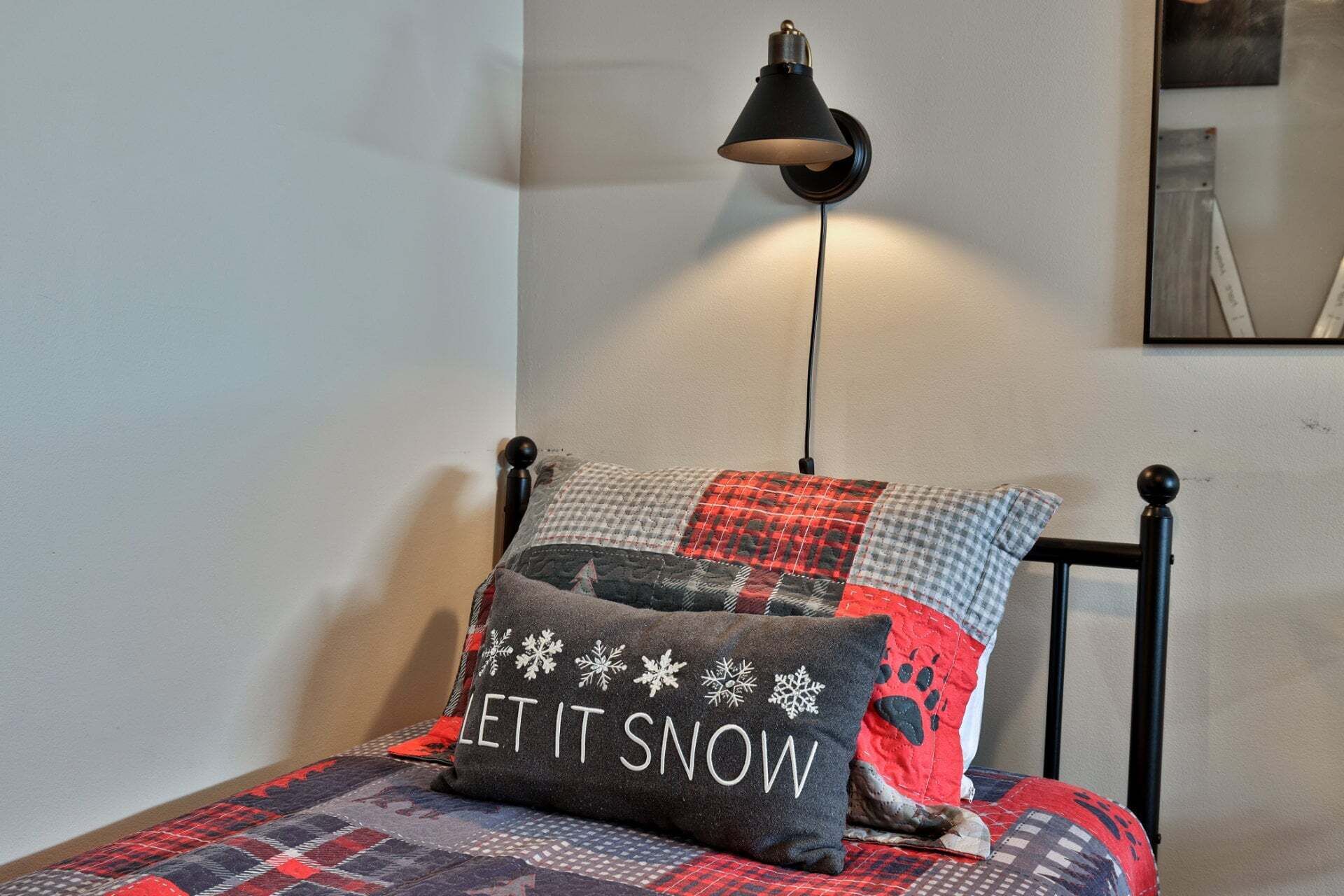 En seng med et tæppe, en pude med teksten 'LET IT SNOW' og en lampe på væggen.
