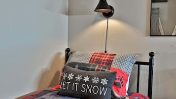 En seng med et tæppe, en pude med teksten 'LET IT SNOW' og en lampe på væggen.