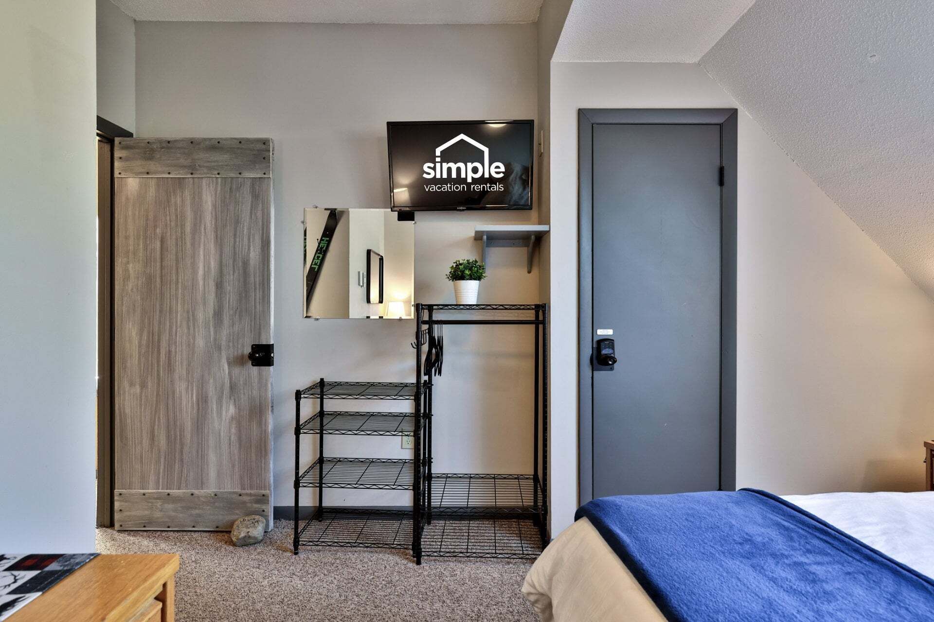 A compact room with a bed, a mounted TV displaying 'simple vacation rentals', a metal shelf with a plant, and a door.