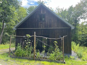 A wooden cabin with a pitched roof, surrounded by a garden with various plants and a fence.
