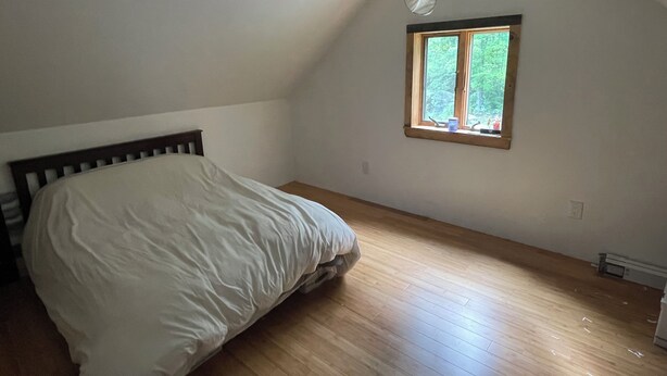Ett sovrum med en enkel säng, trägolv, ett fönster med utsikt över grönområden och ett sluttande tak.