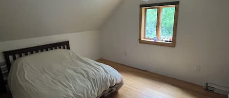 Un dormitorio con una cama individual, suelo de madera, una ventana con vistas a zonas verdes y un techo inclinado.