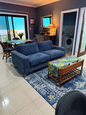 A living room with a blue sofa, a wooden coffee table, and a dining area with a table and chairs. There is a washing machine in the background. - Rivervilla tranquillity by the bay
2 bed unit surrounded by native reserves (Griffin)