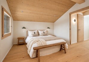 A bedroom with a wooden ceiling, a bed with a wooden bench, a nightstand, and a wall-mounted lamp. - Historic Mazot Retreat by Mrs Miggins – Entire Chalet -Historic Mazot Retreat by Mrs Miggins offers (Lens)