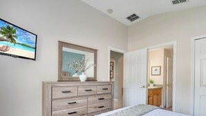 Um quarto com cômoda de madeira, cama com cobertor branco e cinza, televisão fixada na parede e porta que dá para outro espaço.