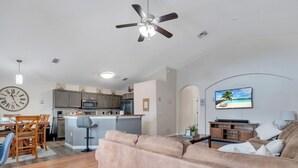 Uma sala moderna com um sofá bege, uma área de jantar com cadeiras de madeira, uma cozinha com armários cinzas e um ventilador de teto.