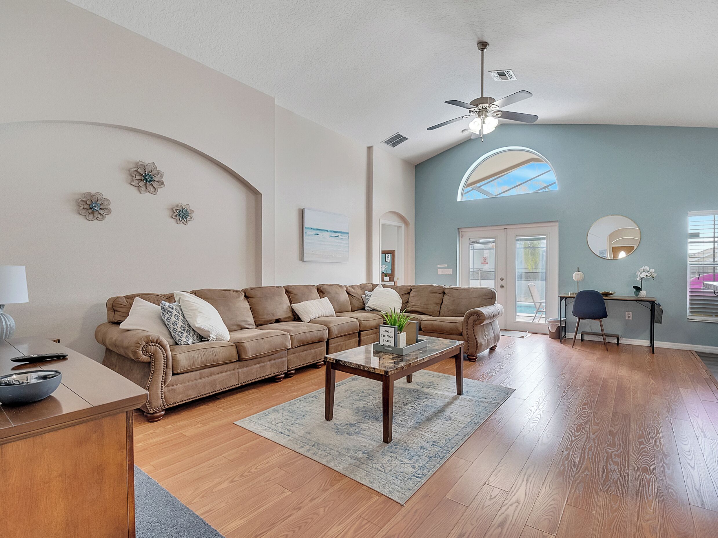 A living room with a brown leather sofa, a coffee table, a ceiling fan, and a view of the outdoors through a large window.