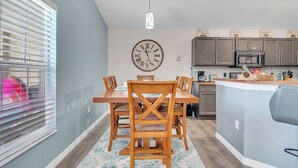Cozinha moderna com um relógio grande, mesa de jantar de madeira e vista para o exterior através de uma janela com persianas.