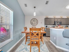 Eine moderne Küche mit einer großen Uhr, einem Holzespeisetisch und Blick ins Freie durch ein Fenster mit Jalousien.