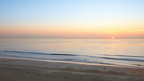 A beach with a calm sea, a visible horizon, and a person walking on the sand.