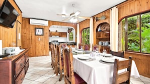 A dining area with a long table set for a meal, wooden chairs, a ceiling fan, and a view of greenery through the windows. - 11 beds, 9 bdrm, 6 bath - Short walk to Bars, Cocal Casino & Beach (Jacó)
