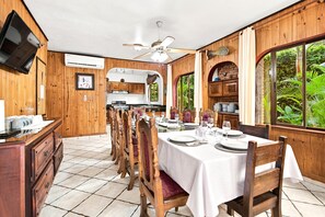 Ein Essbereich mit einem langen Tisch, der für eine Mahlzeit gedeckt ist, Holzstühle, einem Deckenventilator und Blick ins Grüne durch die Fenster.