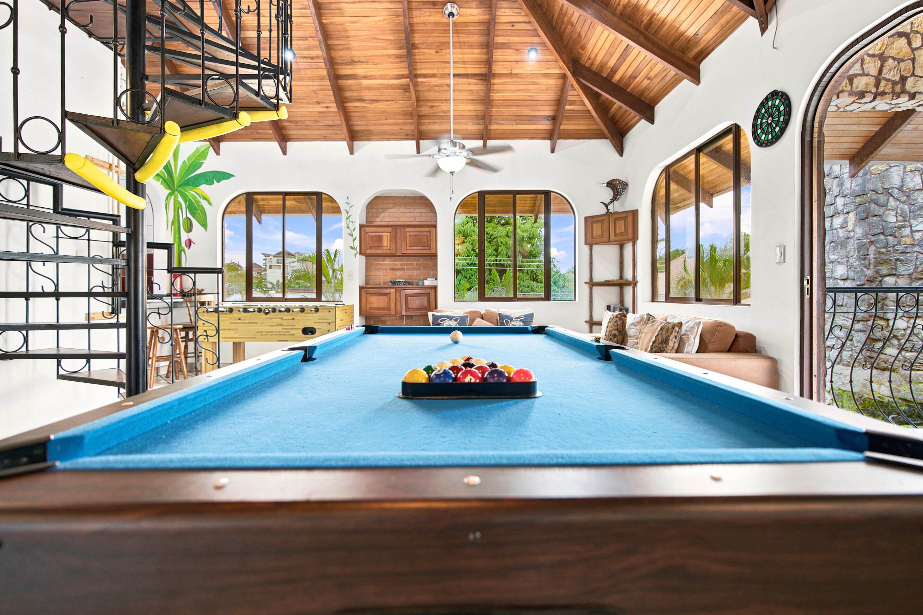 Une table de billard avec les billes au centre, entourée d’un plafond en bois, de fenêtres donnant sur la verdure et d’une porte avec un mur en pierre.
