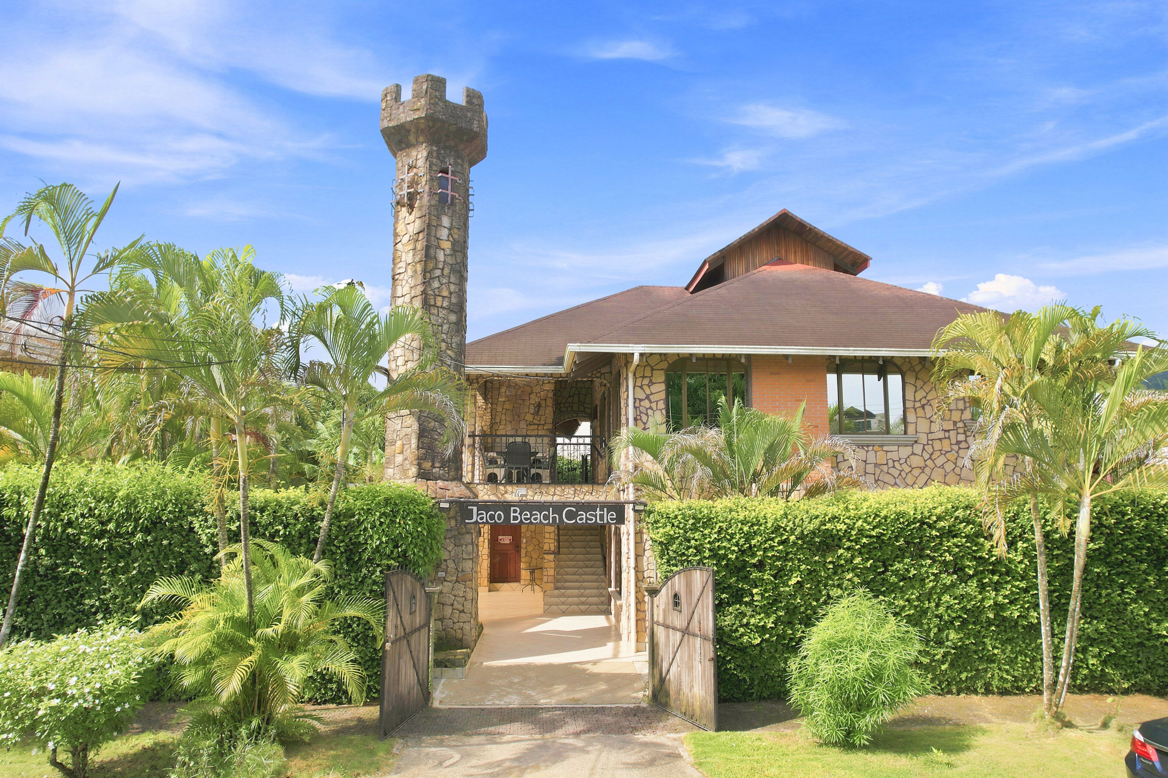 A castle-like building with a stone tower, a covered patio, and lush greenery.