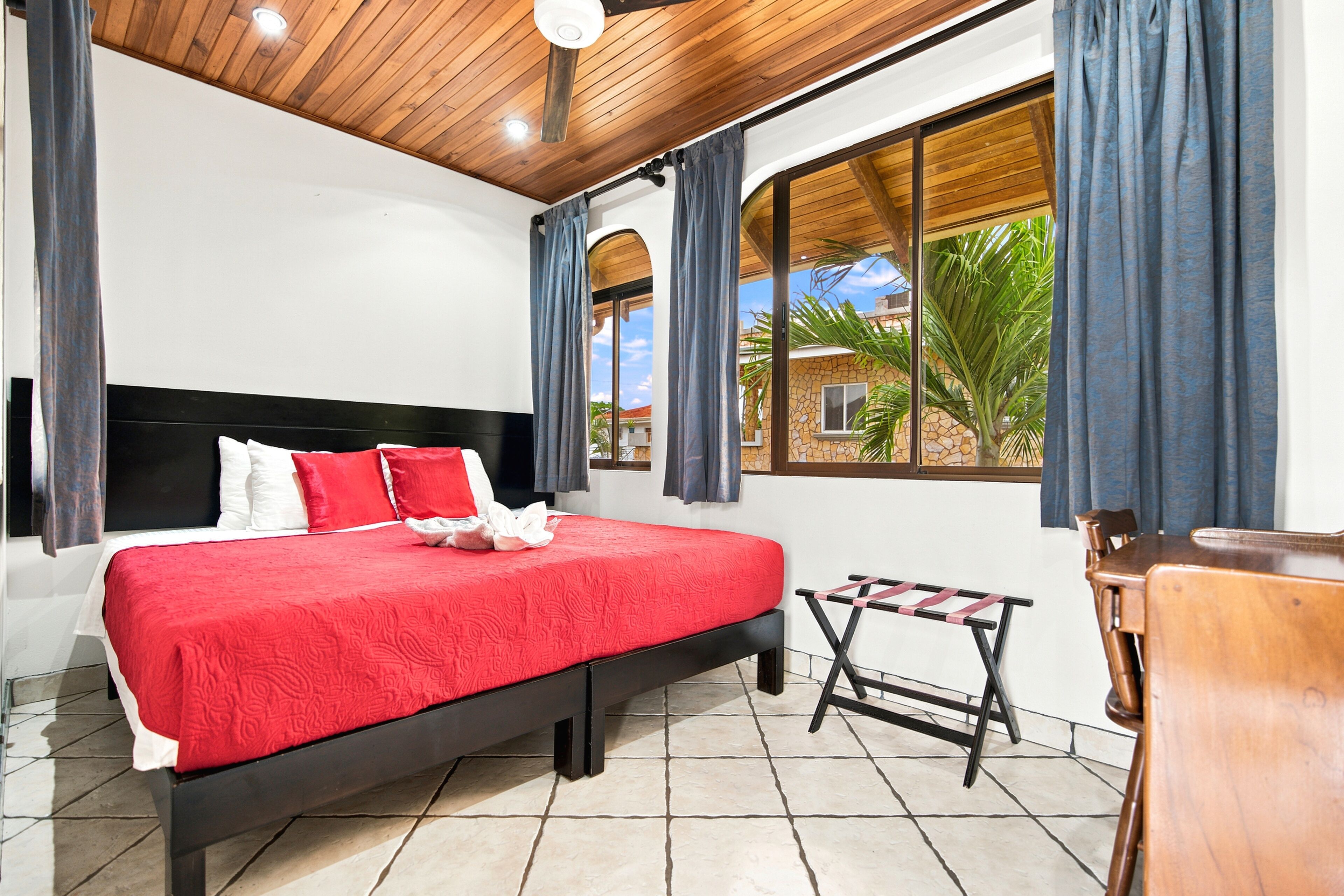 A bedroom with a bed, a window with curtains, a small table, and a chair.