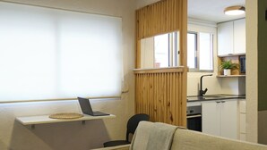 Et kompakt opholdsrum med sofa, et lille bord, en bærbar computer på et stativ, en træskærm og et tekøkken.