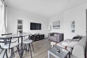 Un salon moderne avec une table ronde en verre, un canapé gris et un téléviseur à écran plat fixé au mur.