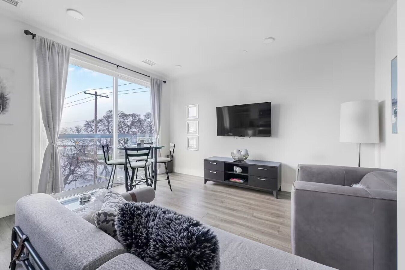 En moderne stue med en grå sofa, et fladskærms-tv, et spisebord med stole og en balkon med udsigt.
