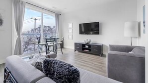 Ein modernes Wohnzimmer mit grauem Sofa, einem Flachbildfernseher, einem Esstisch mit Stühlen und einem Balkon mit Blick.