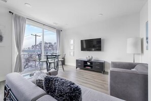 Ein modernes Wohnzimmer mit grauem Sofa, einem Flachbildfernseher, einem Esstisch mit Stühlen und einem Balkon mit Blick.