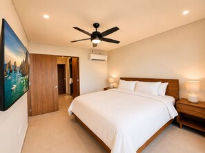 A bedroom with a bed, a ceiling fan, a nightstand, a wall-mounted air conditioner, and a painting of a coastal scene. - CASA CIAN • LUXURY PENTHOUSE SUITE • OCEAN & ARCH VIEWS • PRIVATE ROOFTOP (Cabo San Lucas)