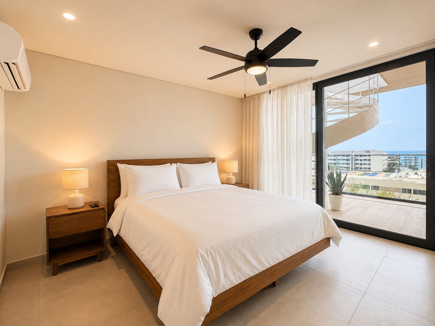 Habitación de hotel con cama, lámparas de noche, ventilador de techo y ventana grande con vistas a edificios y un balcón.