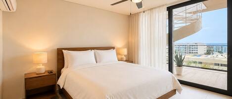 Habitación de hotel con cama, lámparas de noche, ventilador de techo y ventana grande con vistas a edificios y un balcón.