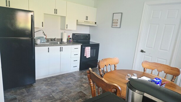 A compact kitchen with white cabinets, a black refrigerator, a microwave, and a dining table with chairs. - Bright & Cozy One Bedroom Getaway (Grand Falls-Windsor)