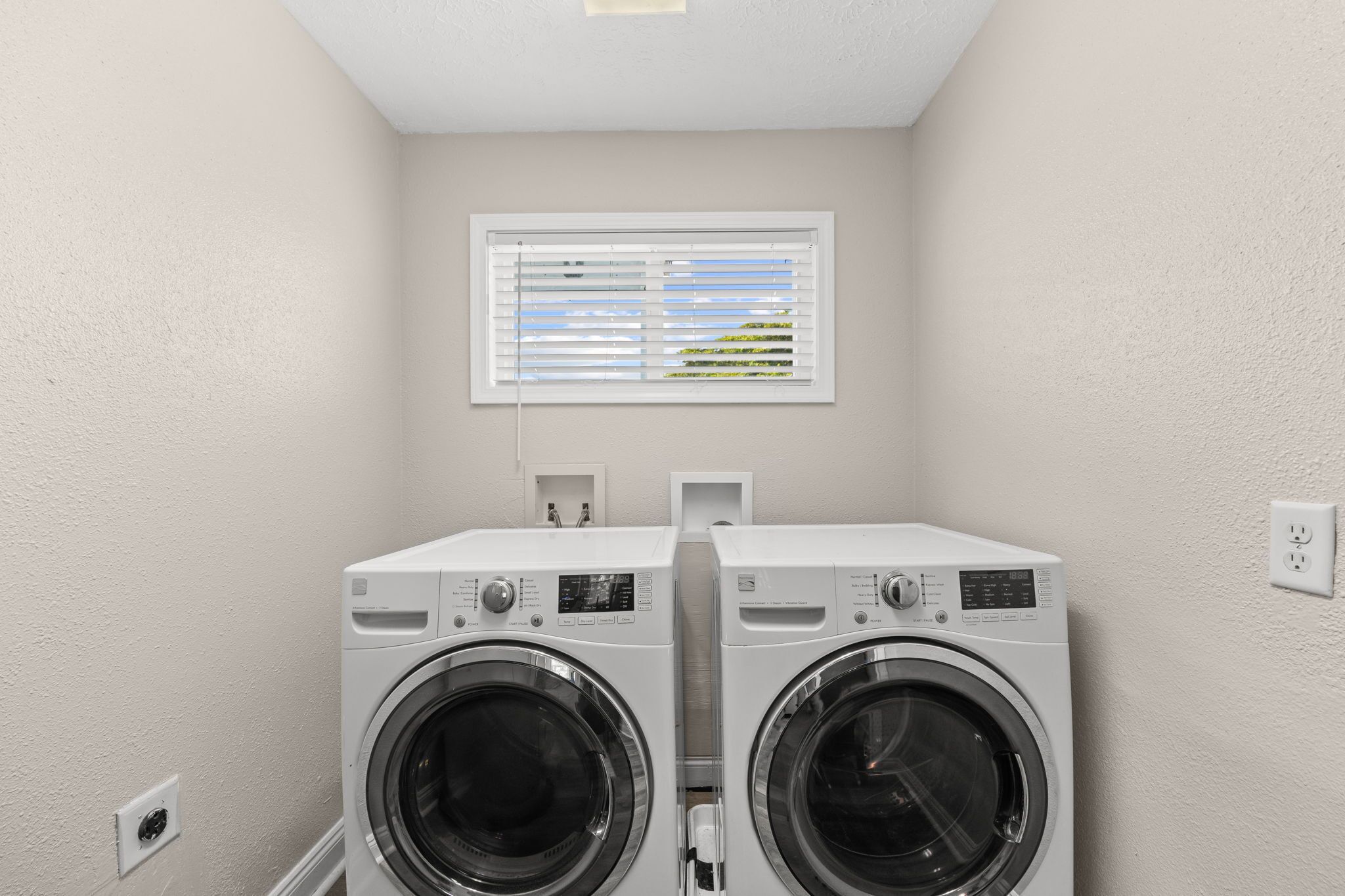A laundry room with two front-loading washing machines, a window with blinds, and a power outlet on the wall.