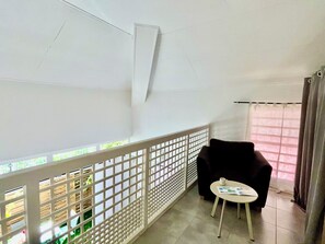 Ein Zimmer mit Balkon, einem kleinen Tisch mit einer Zeitschrift, einem Sessel und einem Fenster mit Vorhängen.