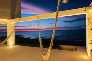 A hammock suspended in a room with a large window overlooking a beach at sunset.