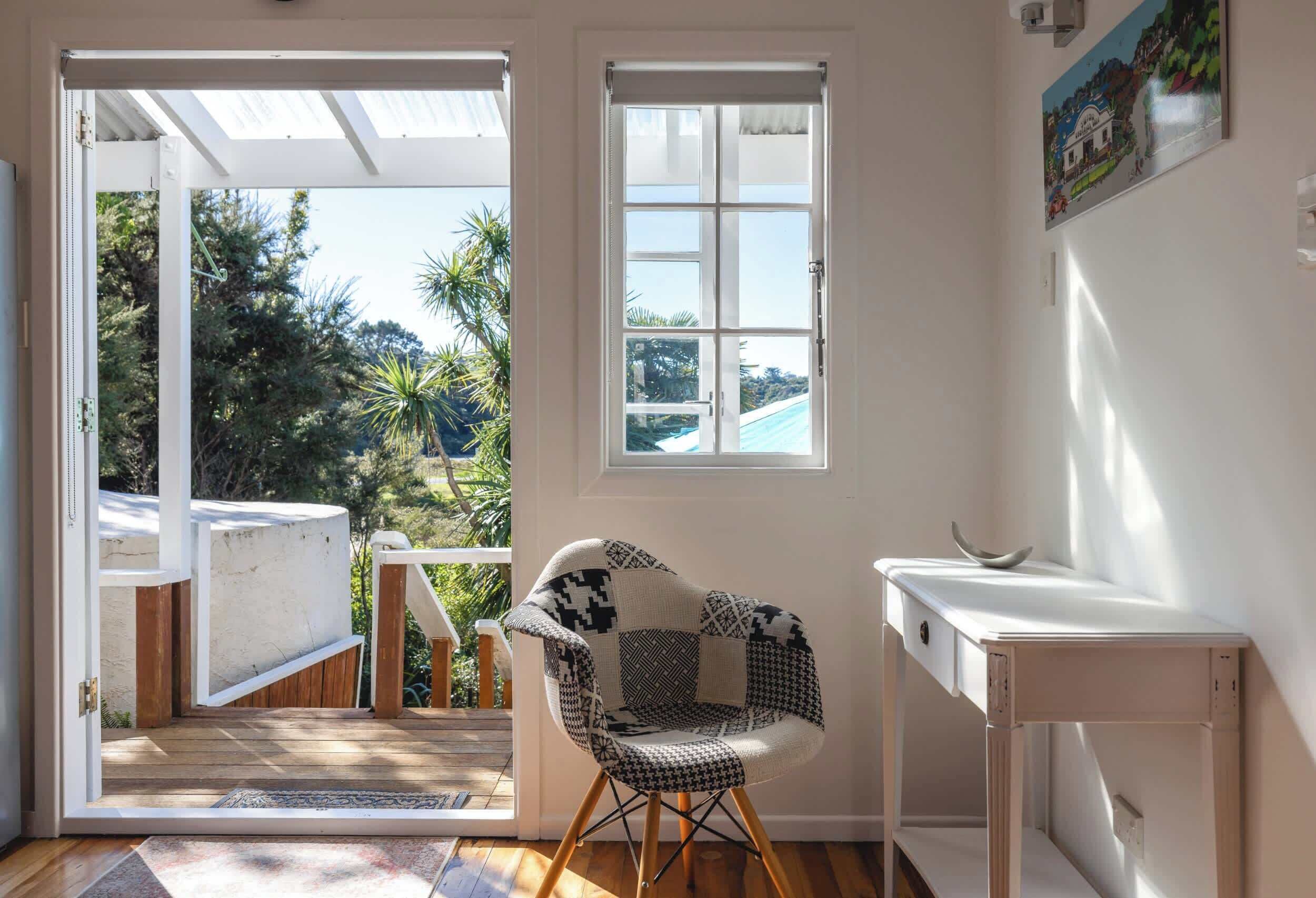 Ein Zimmer mit Blick auf eine Holzterrasse und Grünpflanzen, ein gemustertes Sessel und ein weißer Schreibtisch.