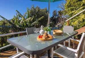 An outdoor dining area with a glass table, chairs, a bottle of Pellegrino water, and a loaf of bread. - Shelly Beach Cottage – Sunny Coastal Stay (Waiheke Island)