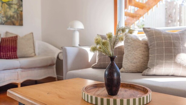 Una sala de estar con una mesa de centro de madera, un jarrón oscuro con plantas secas y un posavasos a rayas.