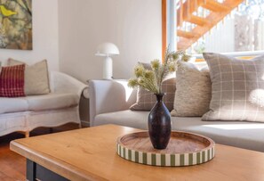 Ein Wohnzimmer mit einem Holztisch, einer dunklen Vase mit getrockneten Pflanzen und einem gestreiften Untersetzer.