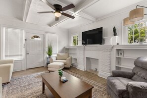 A living room with a fireplace, a ceiling fan, a flat-screen TV, and a large window with a view of greenery. - The Lucile - Spacious 4BR Extended-Stay Home | Fast Wi-Fi & Parking (Wichita Falls)
