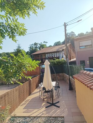 Ein Terrassenbereich im Freien mit Tisch und Stühlen, einem hängenden Sonnenschirm und Blick auf ein Gebäude mit Ziegeldach.