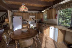 A kitchen with wooden cabinets, a dining table, chairs, a refrigerator, a microwave, a stove, and a window with a view of trees. - Coyote Den | CYH0881 (South Lake Tahoe)