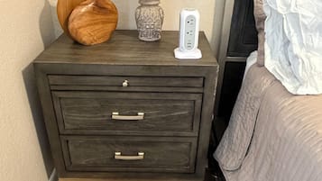 Una mesita de noche de madera oscura con cajones y una lámpara, junto a una cama con un almohadón blanco.