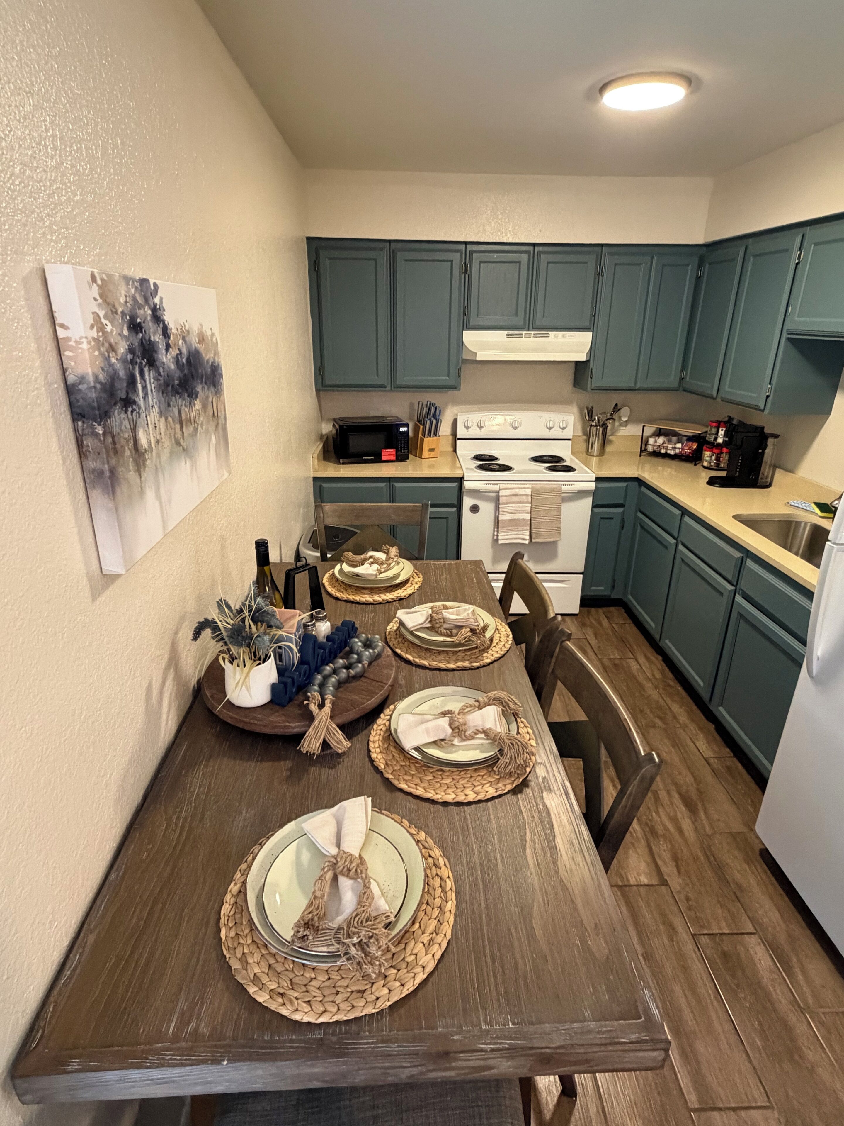 A dining area with a wooden table set for four, a microwave, stove, and cabinets in the background.
