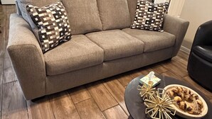 Un salon avec un canapé, deux coussins à motifs, une table basse agrémentée d’objets décoratifs, et une cuisine en arrière-plan.