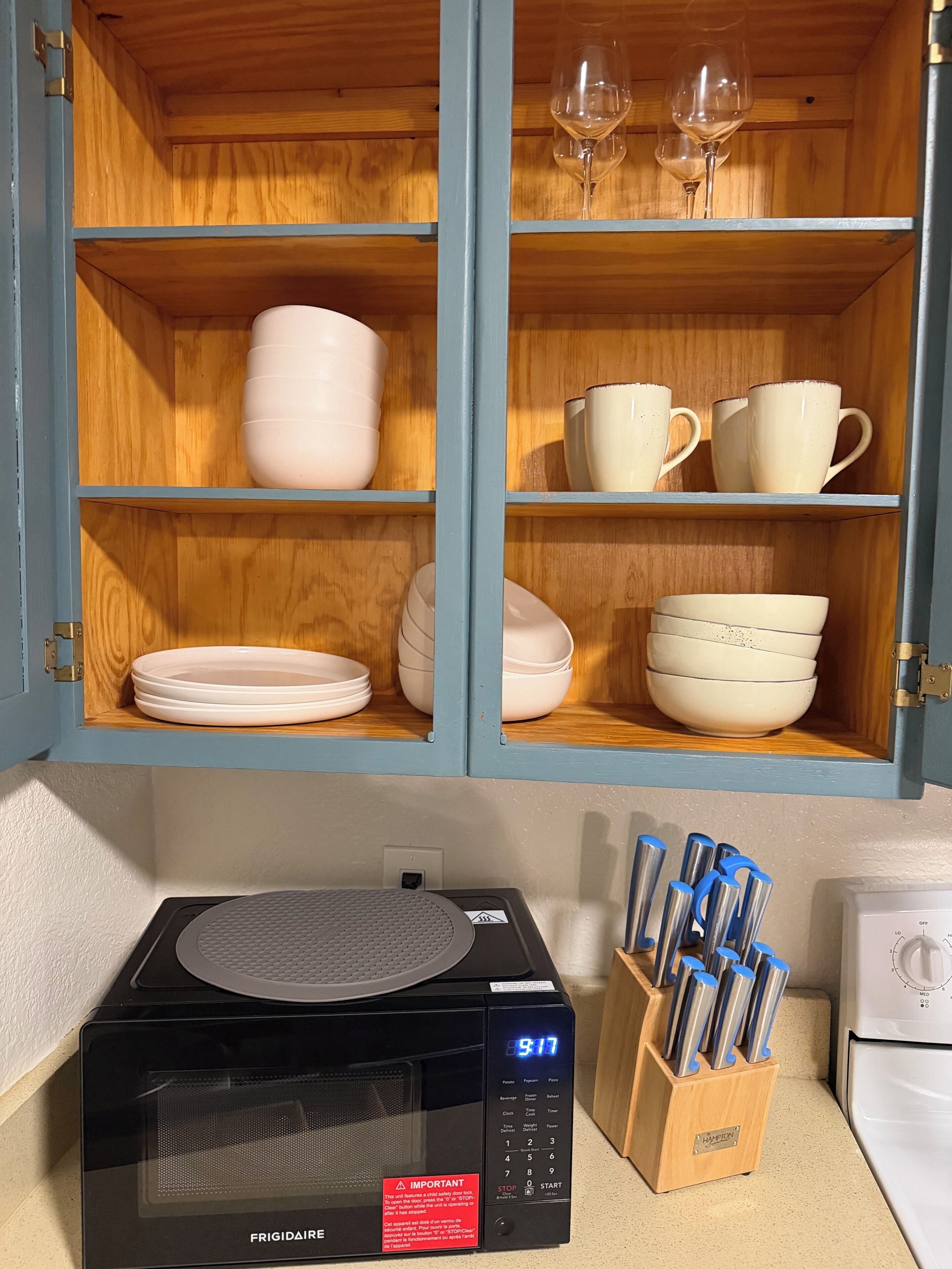 Una cocina con microondas, bloque de cuchillos y un armario con vajilla y vasos.