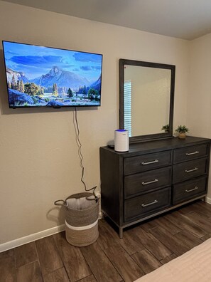Una habitación con una televisión instalada en la pared mostrando un paisaje pintoresco, un aparador de madera oscura con cajones, un espejo encima y una cesta de mimbre en el suelo.