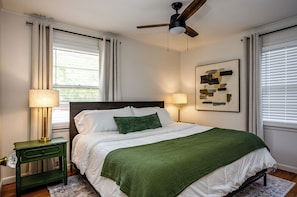 A bedroom with a bed, two bedside lamps, a ceiling fan, and a painting on the wall.