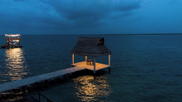 A thatched-roof structure with a table and chairs on a floating platform in the water. - GlampCalar (Bacalar)