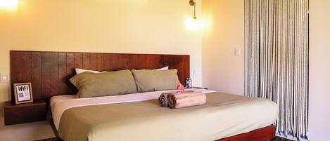 A hotel room with a wooden bed, a wall-mounted air conditioner, a bedside table with a framed item, and a curtain.
