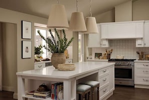 Een keuken met witte kasten, een centraal eiland met krukken, hangende pendelverlichting en een fornuis.